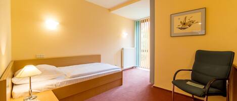 A hotel room with a single bed, a chair, a bedside table with a lamp and phone, and a framed picture on the wall.