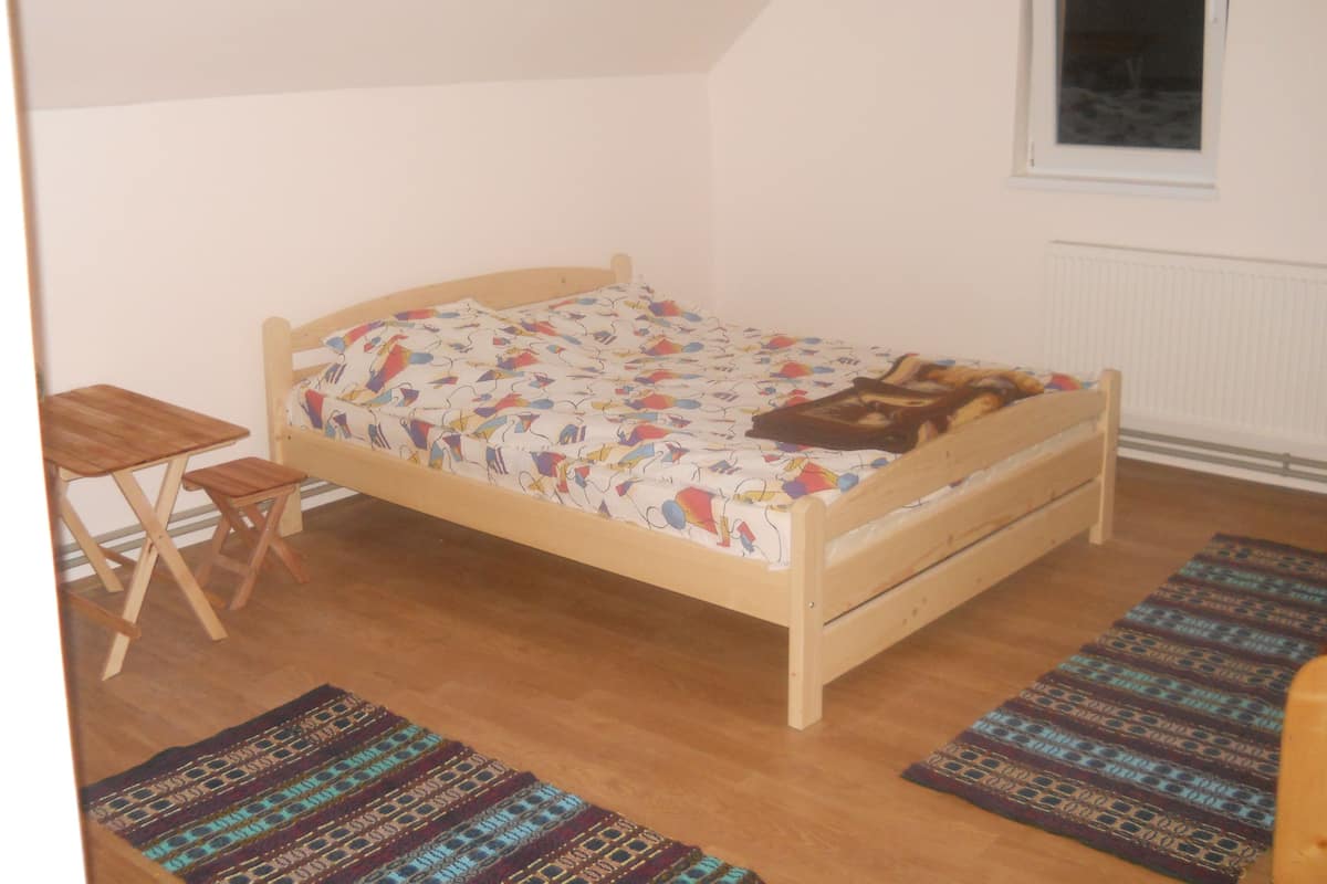A single bed with a colorful bedspread, a small wooden table, and two rugs on a wooden floor in a room with white walls and a window.