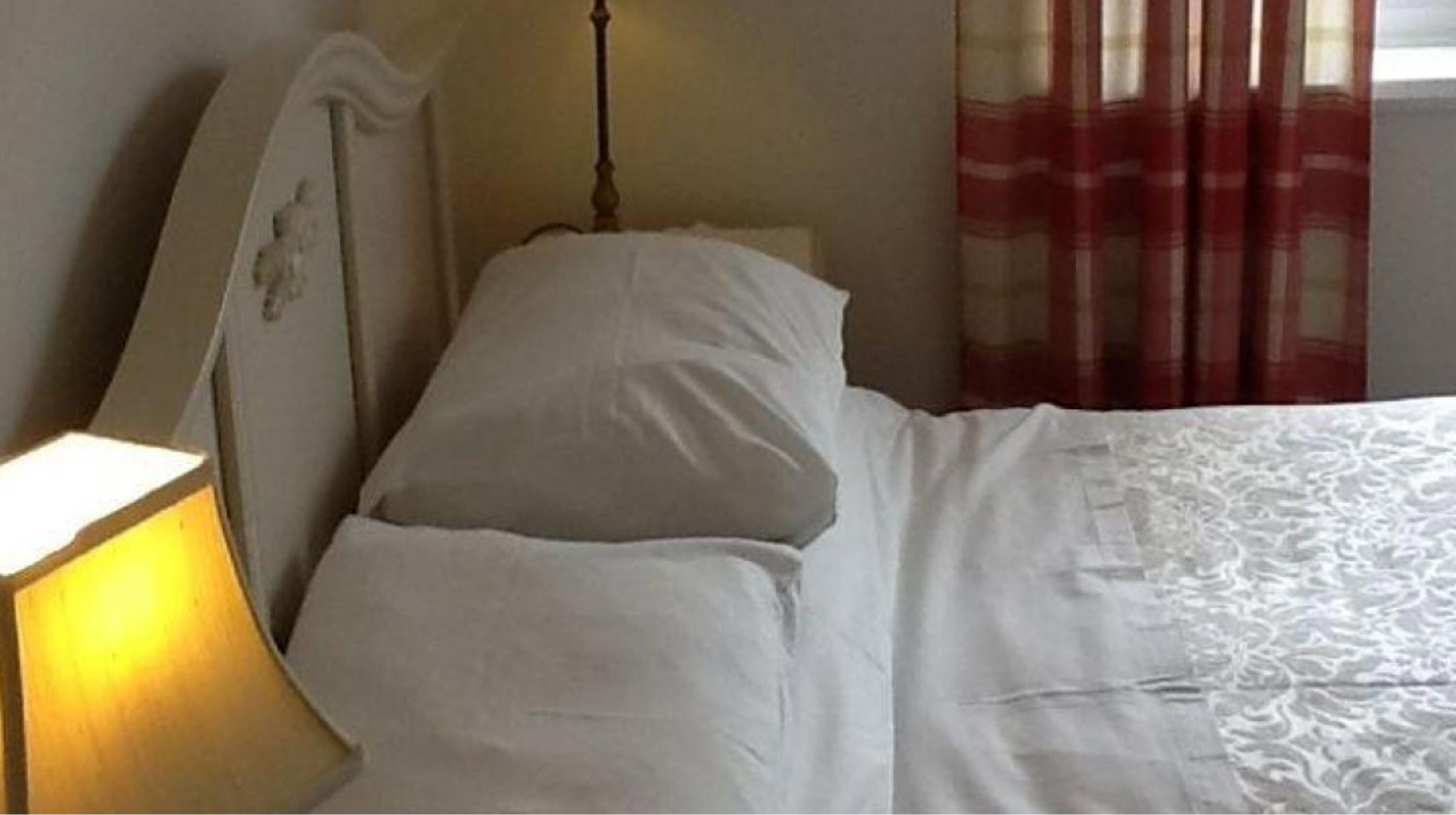 Una cama con ropa blanca, una lámpara de noche y una ventana con cortinas rojas.
