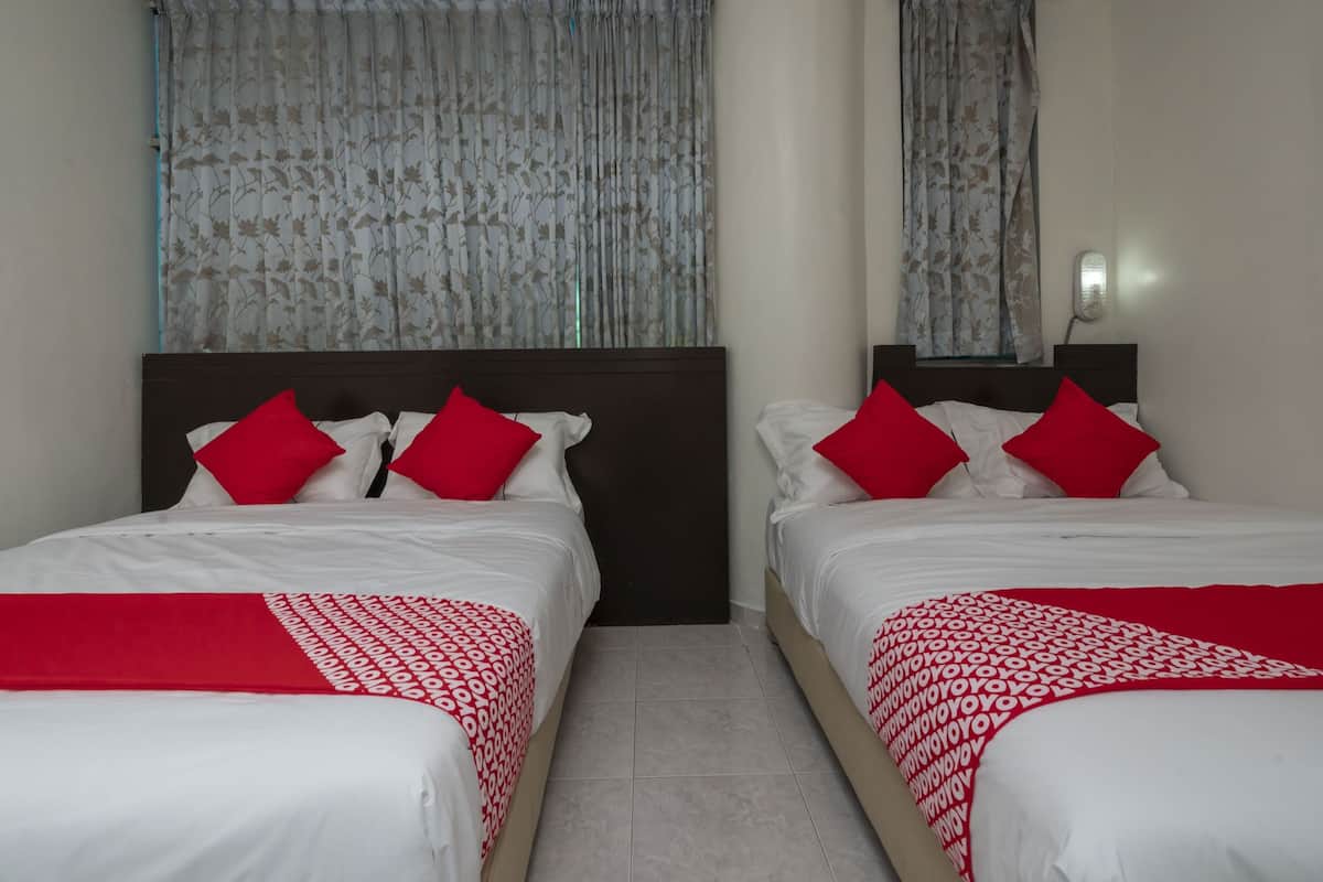 A hotel room with two beds, each with white bedding and red pillows. The room has a plain white wall and a window with floral curtains.