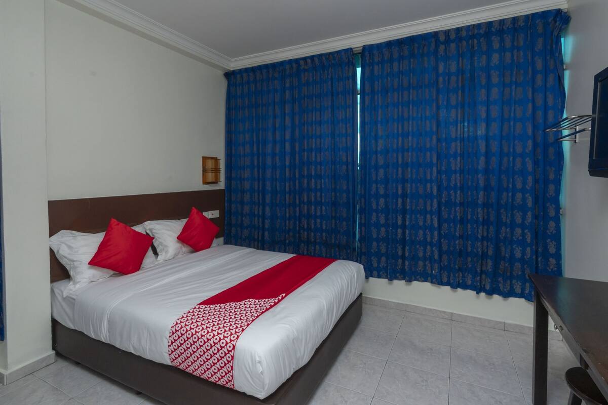 A hotel room with a bed, red and white bedding, a blue curtain, and a wooden nightstand.