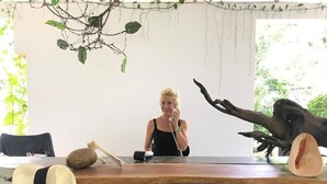 A person sitting at a wooden table with a microphone, surrounded by decorative items like a hat and a shell. - Five (Morakatiyara)