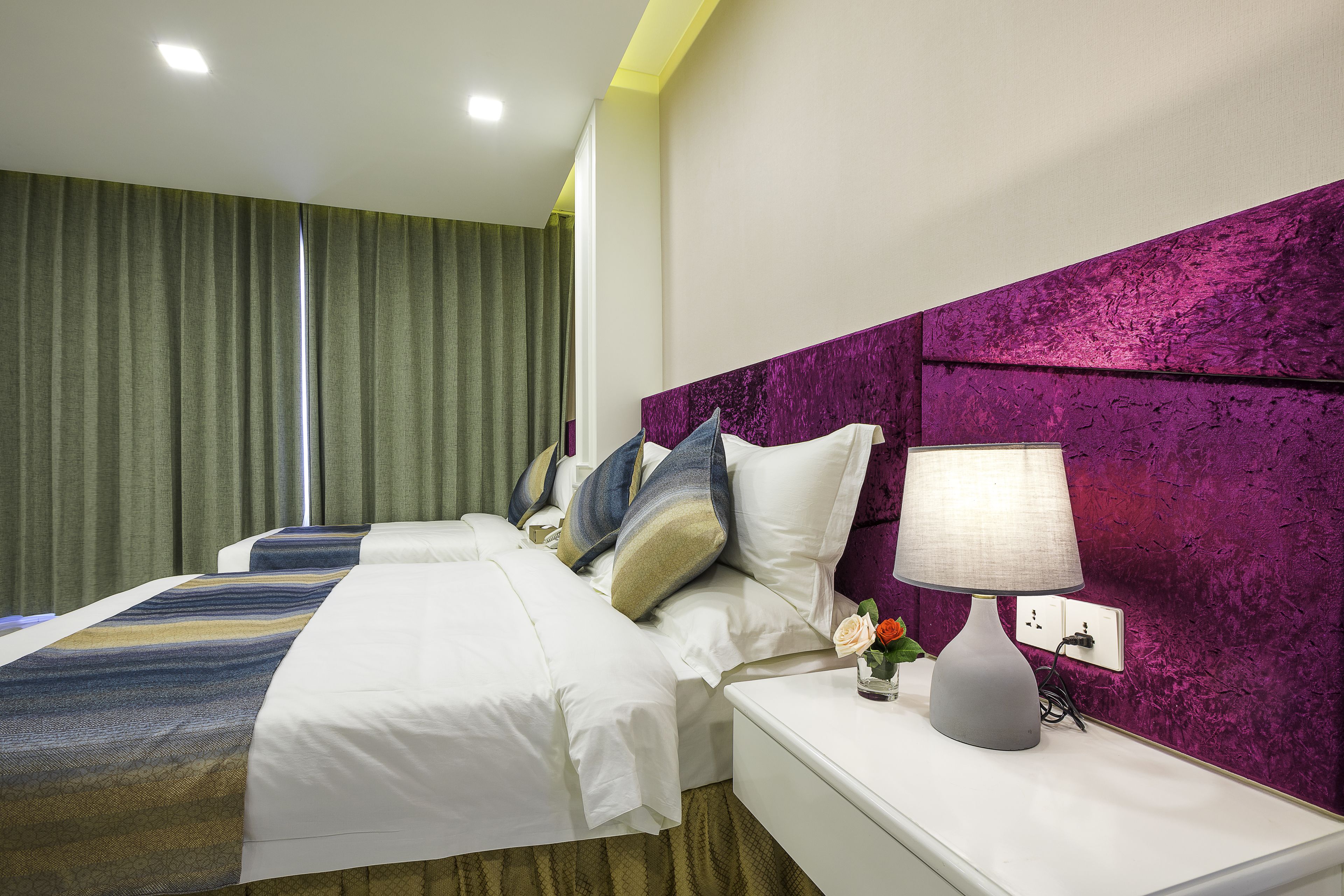 A hotel room with two beds, a purple headboard, a bedside table with a lamp, and a green curtain.