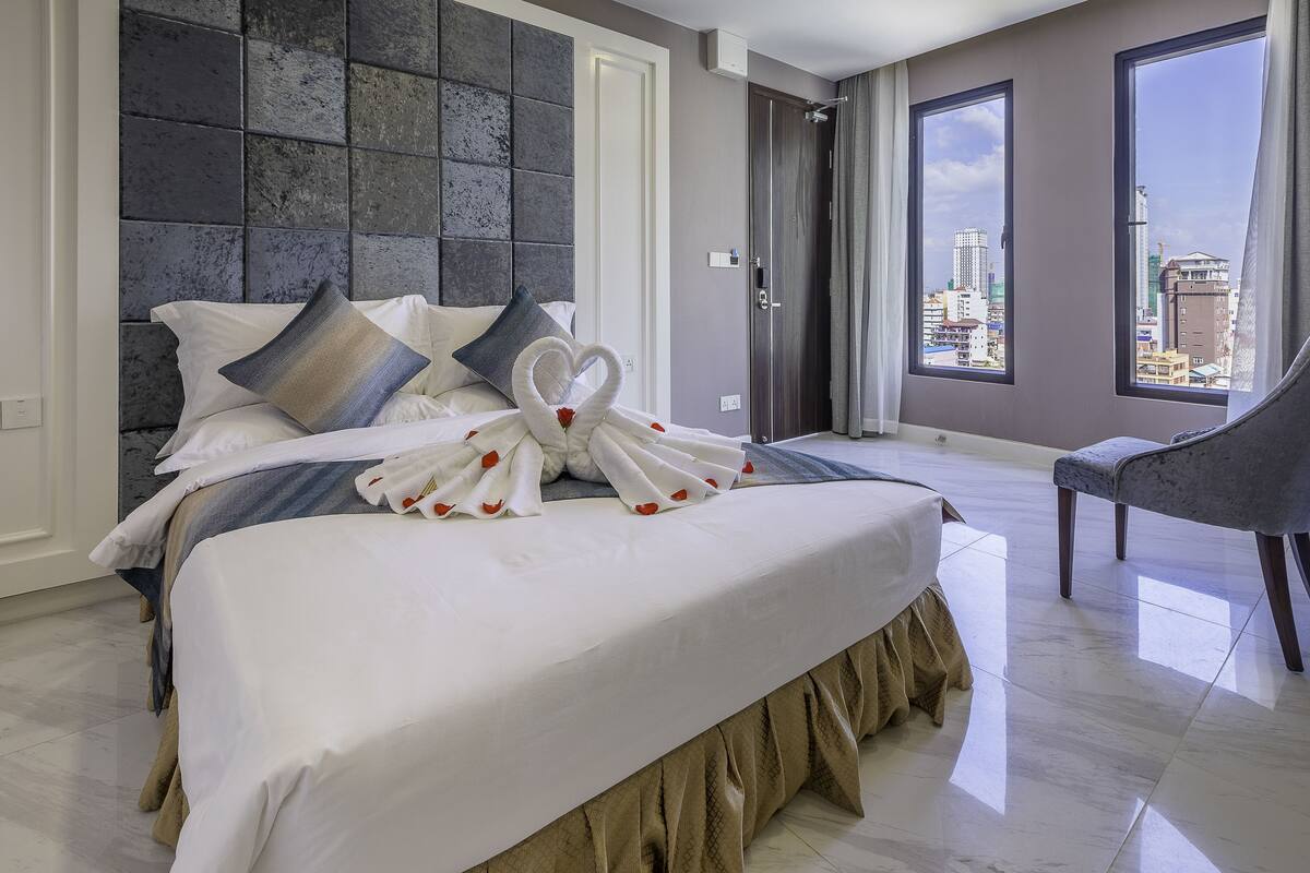 A modern hotel room with a large bed, a city view, and a grey armchair.