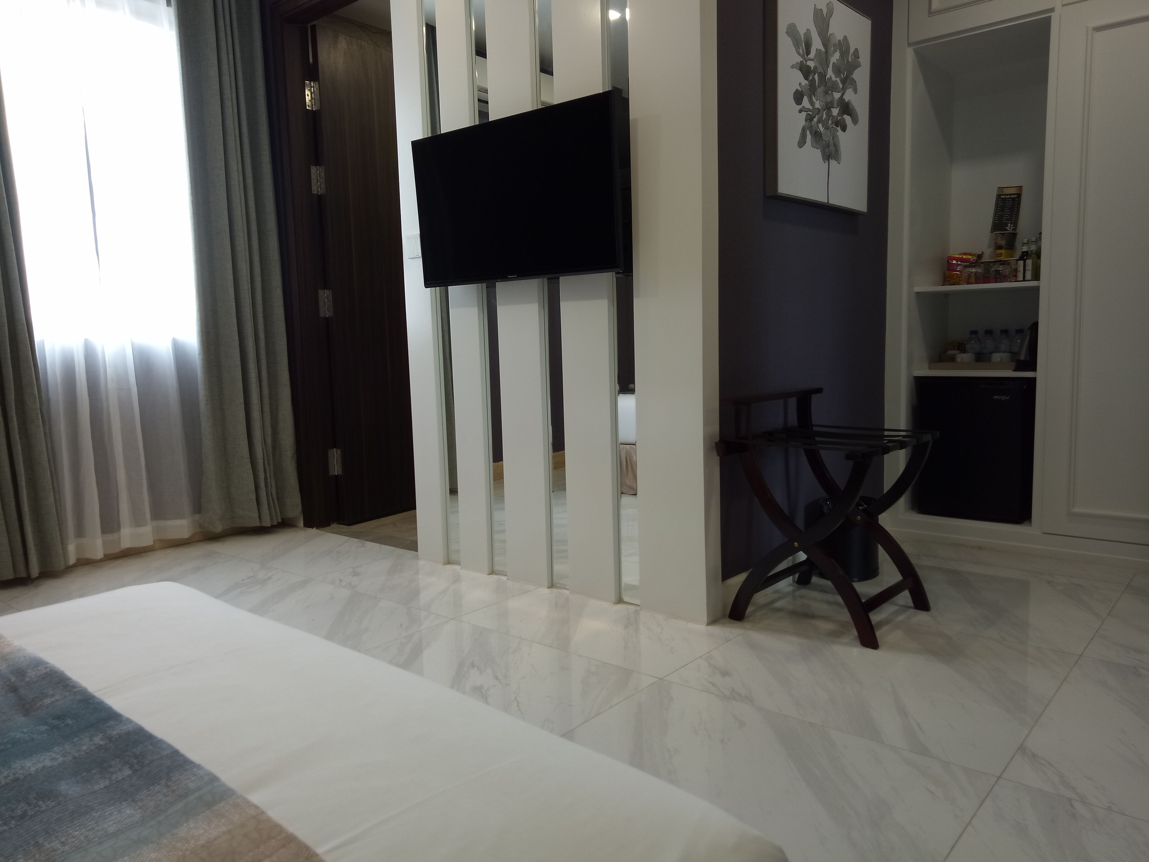 A modern living room with a flat-screen TV mounted on a wall, a dining area with a table and chairs, and a kitchen area with a refrigerator and shelves.