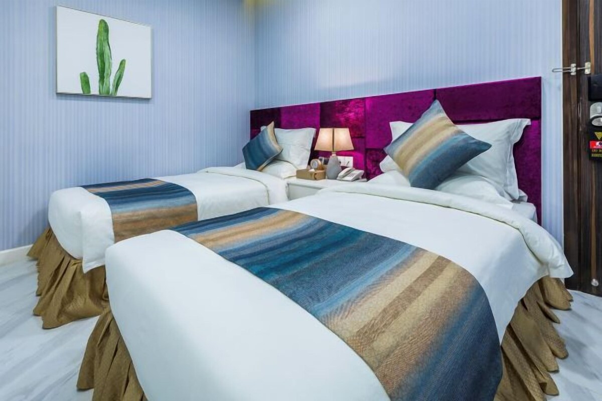 Une chambre d’hôtel avec deux lits, une tête de lit violette, une table de chevet avec une lampe et une œuvre d’art représentant un cactus accrochée au mur.