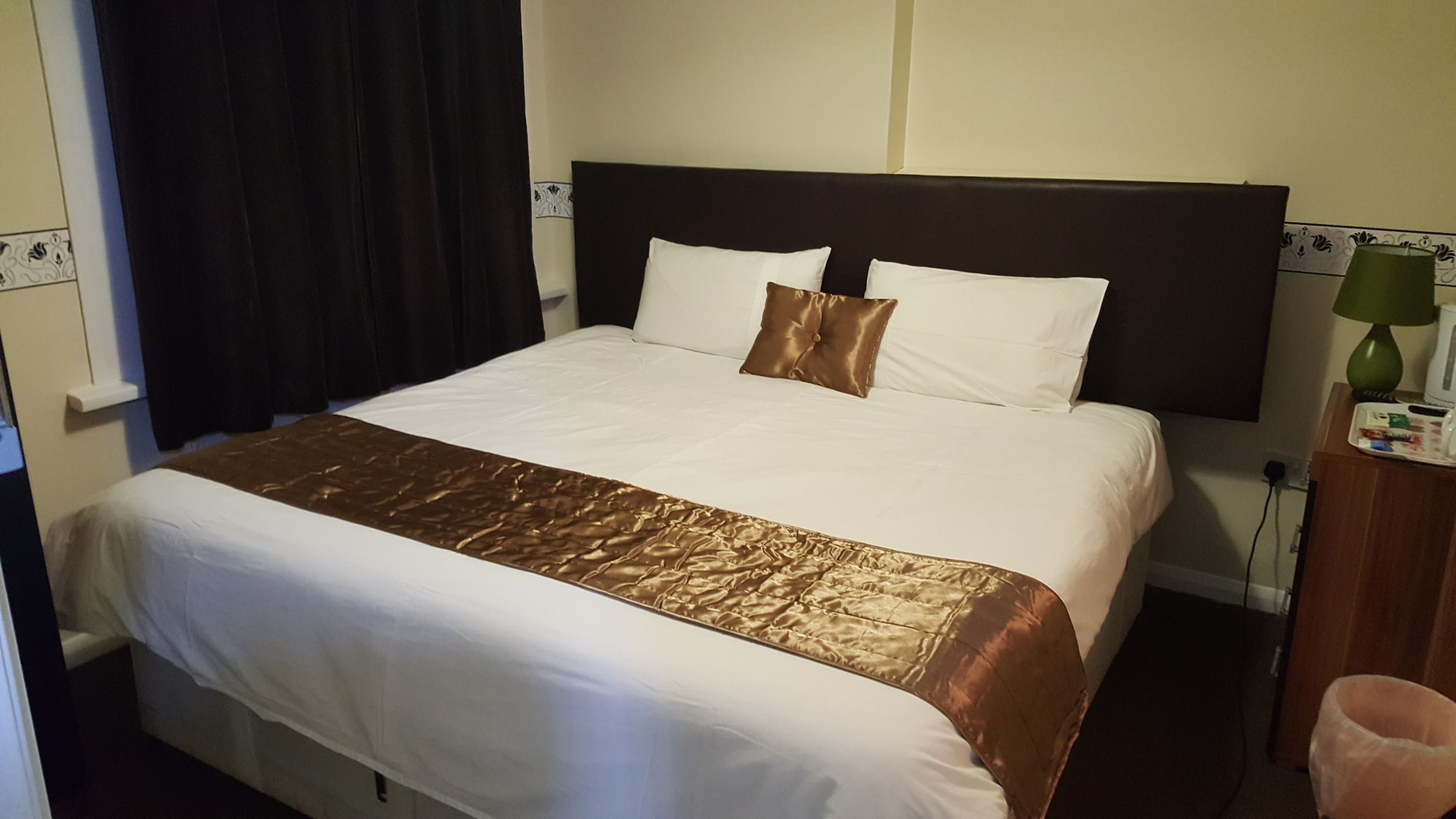A hotel room with a large bed, a brown bedspread, white pillows, and a green lamp on a wooden nightstand.