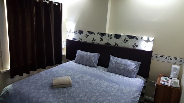 A hotel room with a bed, a nightstand, a window with curtains, and a wall with a floral pattern.