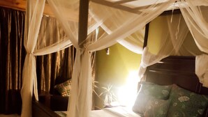 Una cama con dosel y cuatro columnas, con una colcha verde estampada, cabecera de madera y una mesita de noche con lámpara.