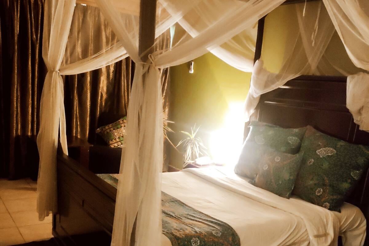 Un lit à baldaquin à quatre poteaux, avec une couvre-lit à motifs verts, une tête de lit en bois et une table de chevet avec une lampe.