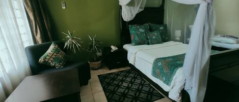A bedroom with a bed under a canopy, a desk, two potted plants, and a patterned rug.