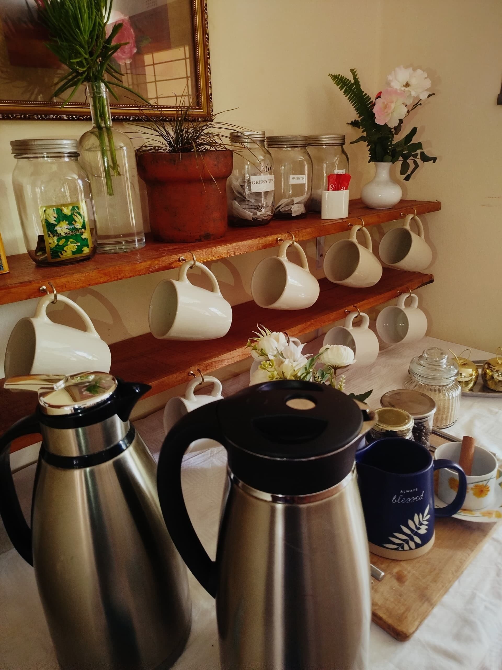 Een kitchenette met roestvrijstalen koffiezetapparaten, witte mokken op planken en diverse potten en bakken.