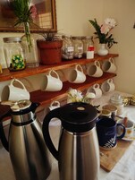A kitchen area with stainless steel coffee makers, white mugs on shelves, and a variety of jars and containers.