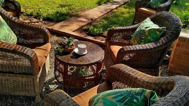 Un área de estar al aire libre con mobiliario de mimbre y una pequeña mesa, rodeada por un césped y un jardín bien cuidados.