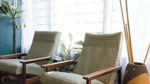 Dos sillones con tapicería estampada, una mesita de madera, un jarrón decorativo con ramas y un ventanal con cortinas translúcidas.