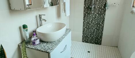 Een badkamer met een wit wastafelmeubel, een spiegel, een douche met een muur van kiezelstenen, een toilet en een vloerkleed met een patroon.