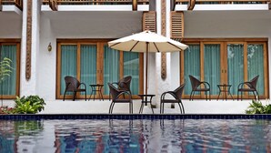 Un área de piscina con una mesa y sillas, rodeada de ventanas con contraventanas de madera, y una fachada de edificio blanca.