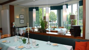A dining area with tables set for a meal, a green upholstered bench, and a view of the outdoors through large windows. - Hotel Landgasthof Evering (Emsbueren)