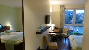 A hotel room with a bed, a desk, a chair, a sofa, a TV, and a window with a view of houses. - Hotel Landgasthof Evering (Emsbueren)