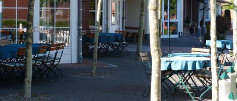 An outdoor seating area with tables and chairs, a brick building, and a tree in the foreground.