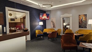 Vestíbulo de hotel con recepción, área de descanso con sillas amarillas y azules, televisor mostrando un fuego y obras de arte en las paredes.