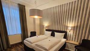 Cama doble con dos almohadas, una mesita de noche, una silla, una lámpara y una ventana con cortinas.