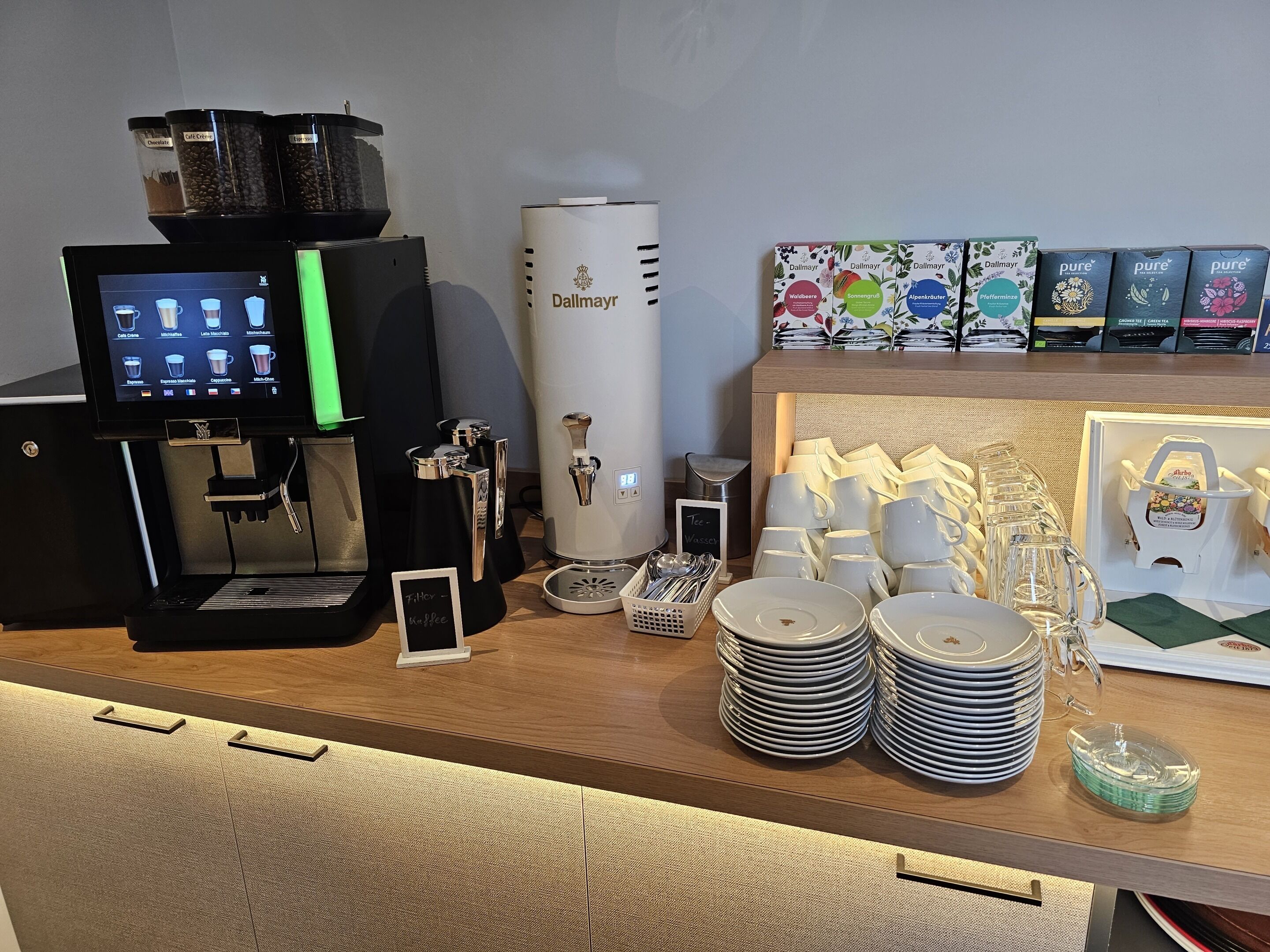 A coffee station with a coffee machine, a milk frother, and a coffee grinder. There are stacks of plates and cups, and a display of coffee pods.