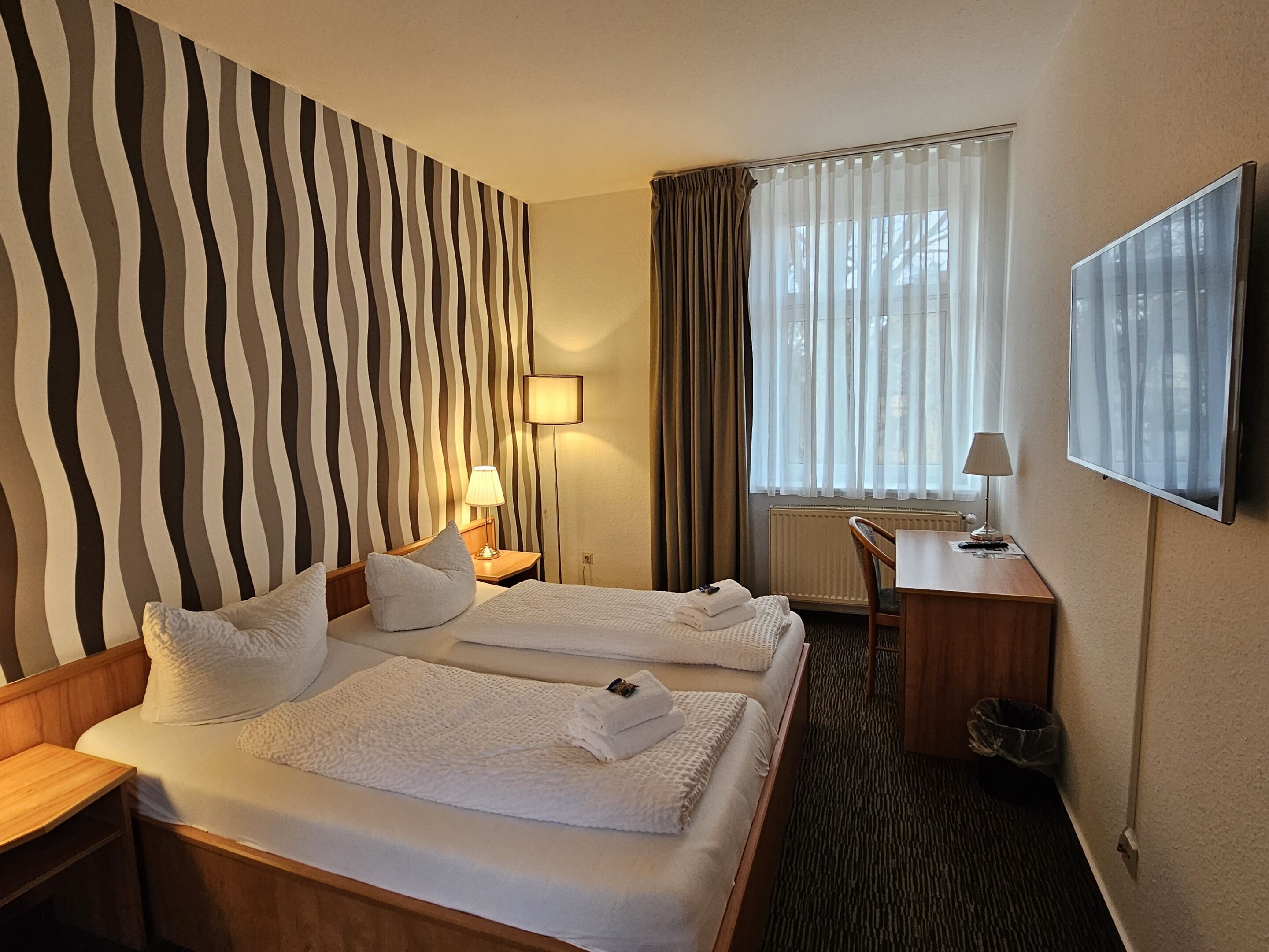 Ein Hotelzimmer mit zwei Betten, einem Schreibtisch, einem Stuhl, einem Fernseher und einem Fenster mit Vorhängen.