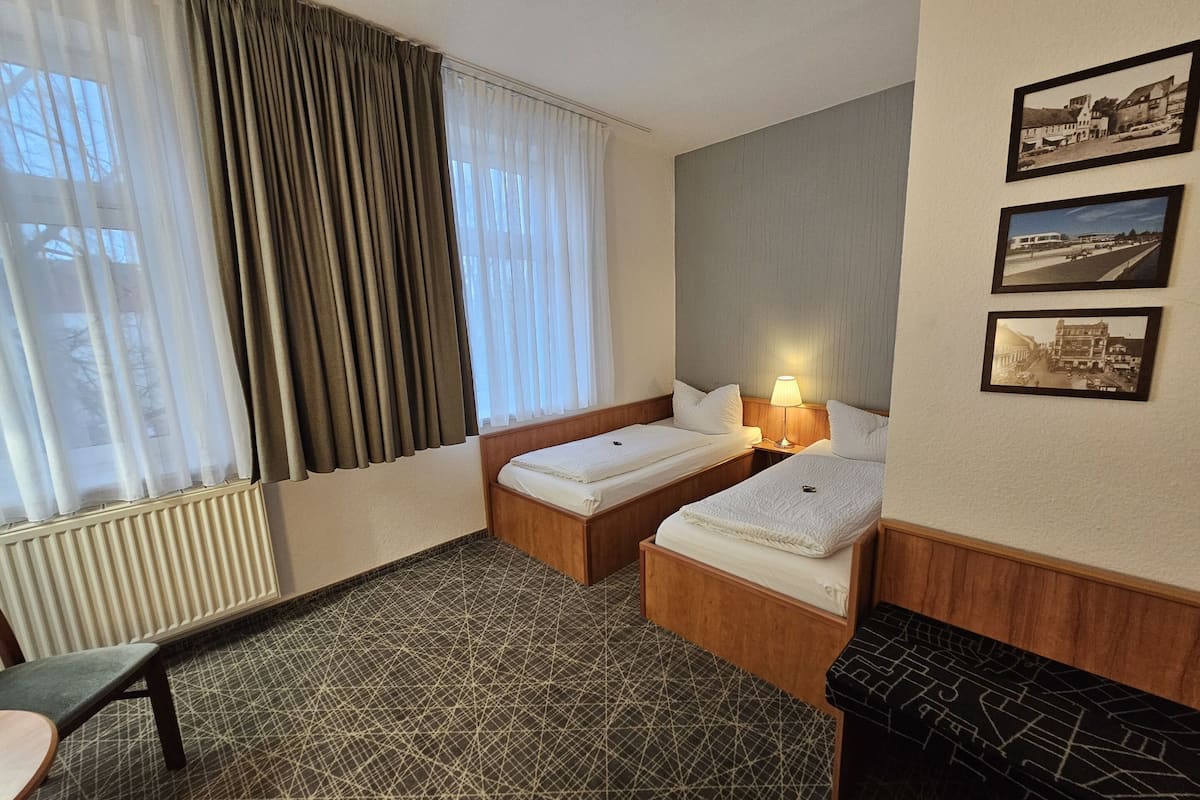 Ein Hotelzimmer mit zwei Betten, einem Heizkörper, einem Schreibtisch und einem Stuhl. Vor den Fenstern hängen Vorhänge und an der Wand befinden sich drei gerahmte Bilder.