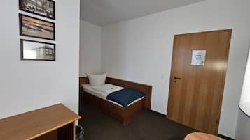 A small, single-bed room with a wooden bed, a desk, and a chair. There are three framed pictures on the wall.