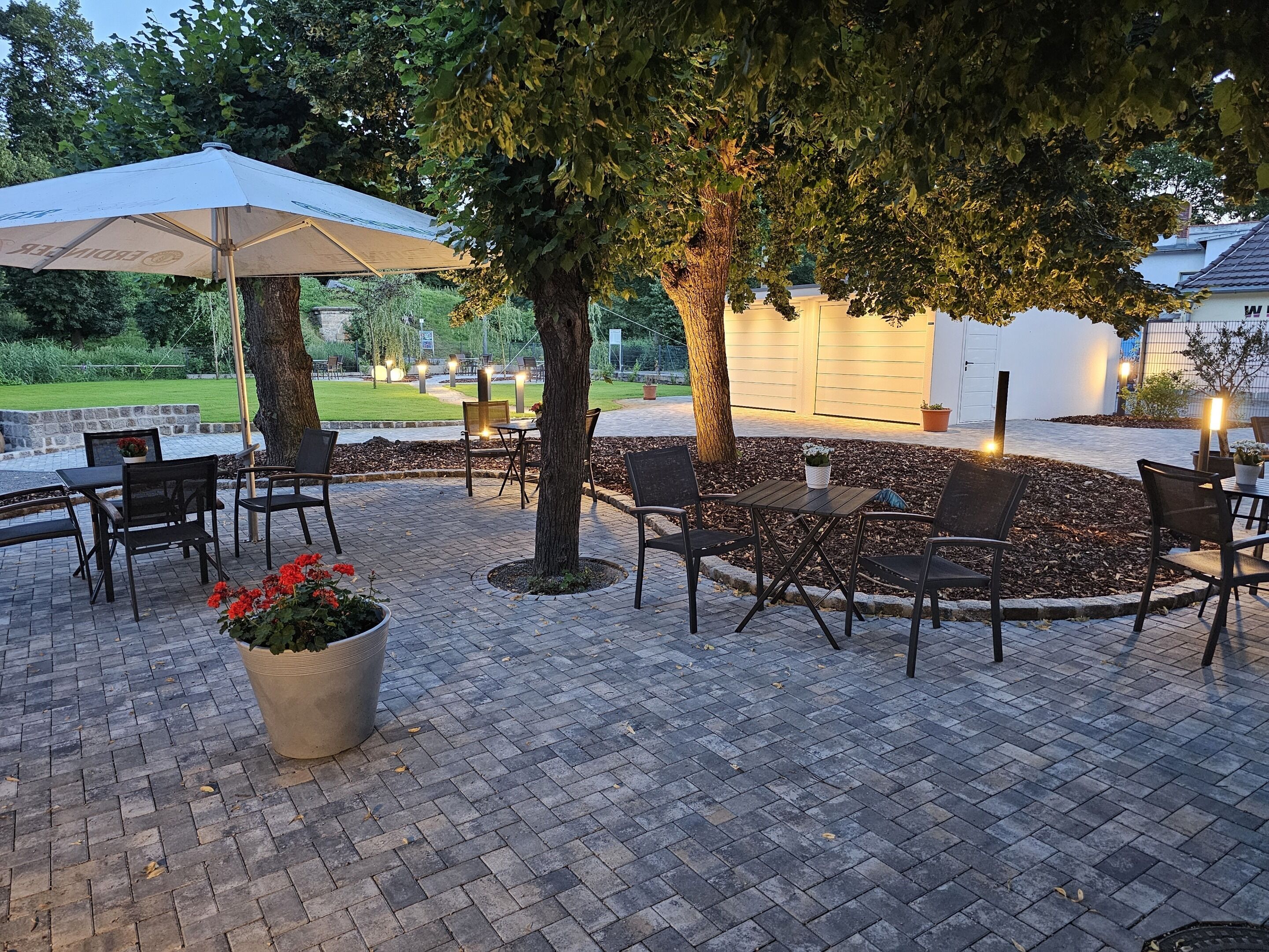 Un patio al aire libre con suelo de ladrillo, varias mesas y sillas, un paraguas grande y plantas en macetas.