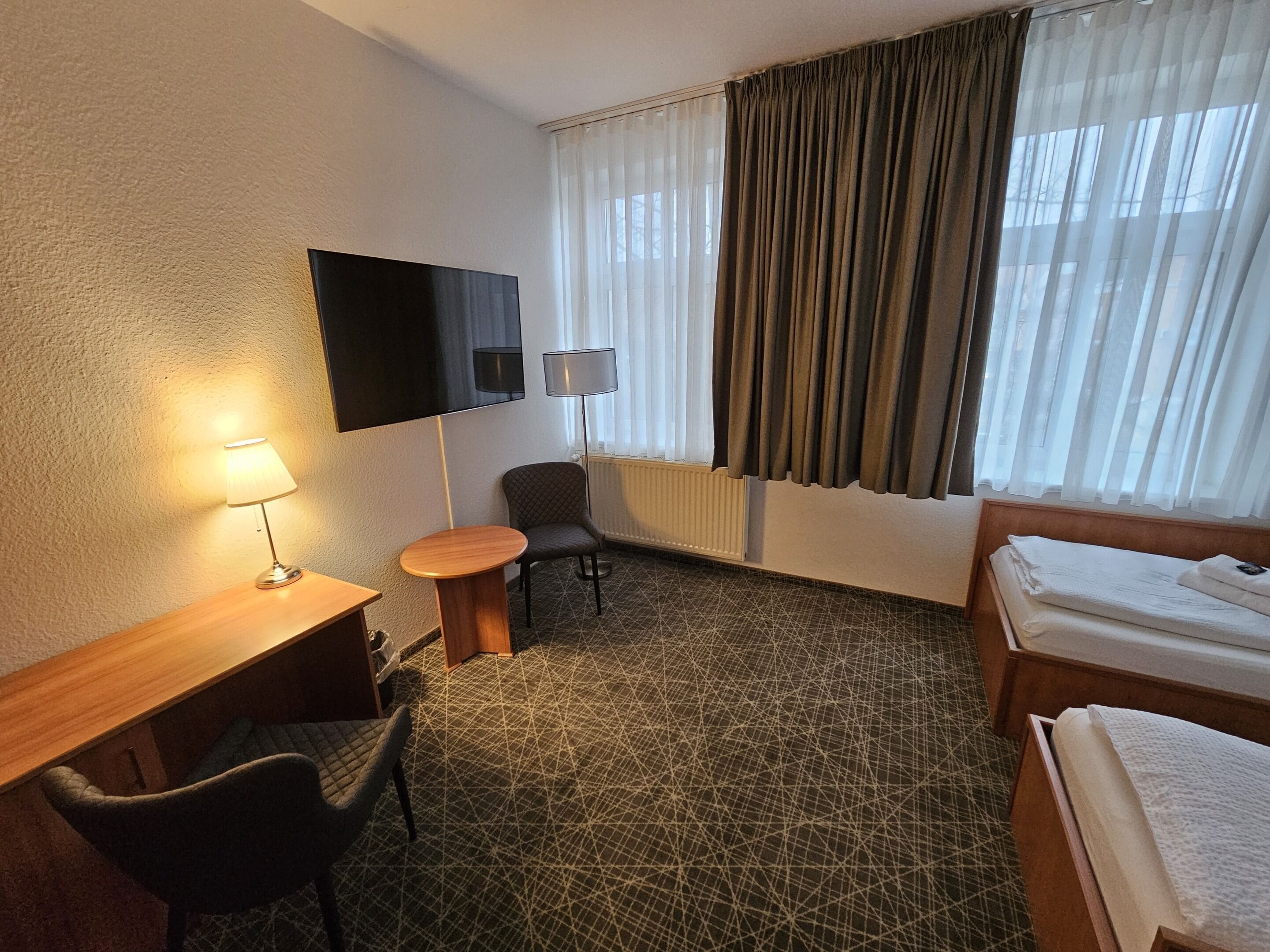 Ein Hotelzimmer mit einem Bett, einem Schreibtisch mit Lampe, einem Sessel und einem an der Wand befestigten Fernseher.