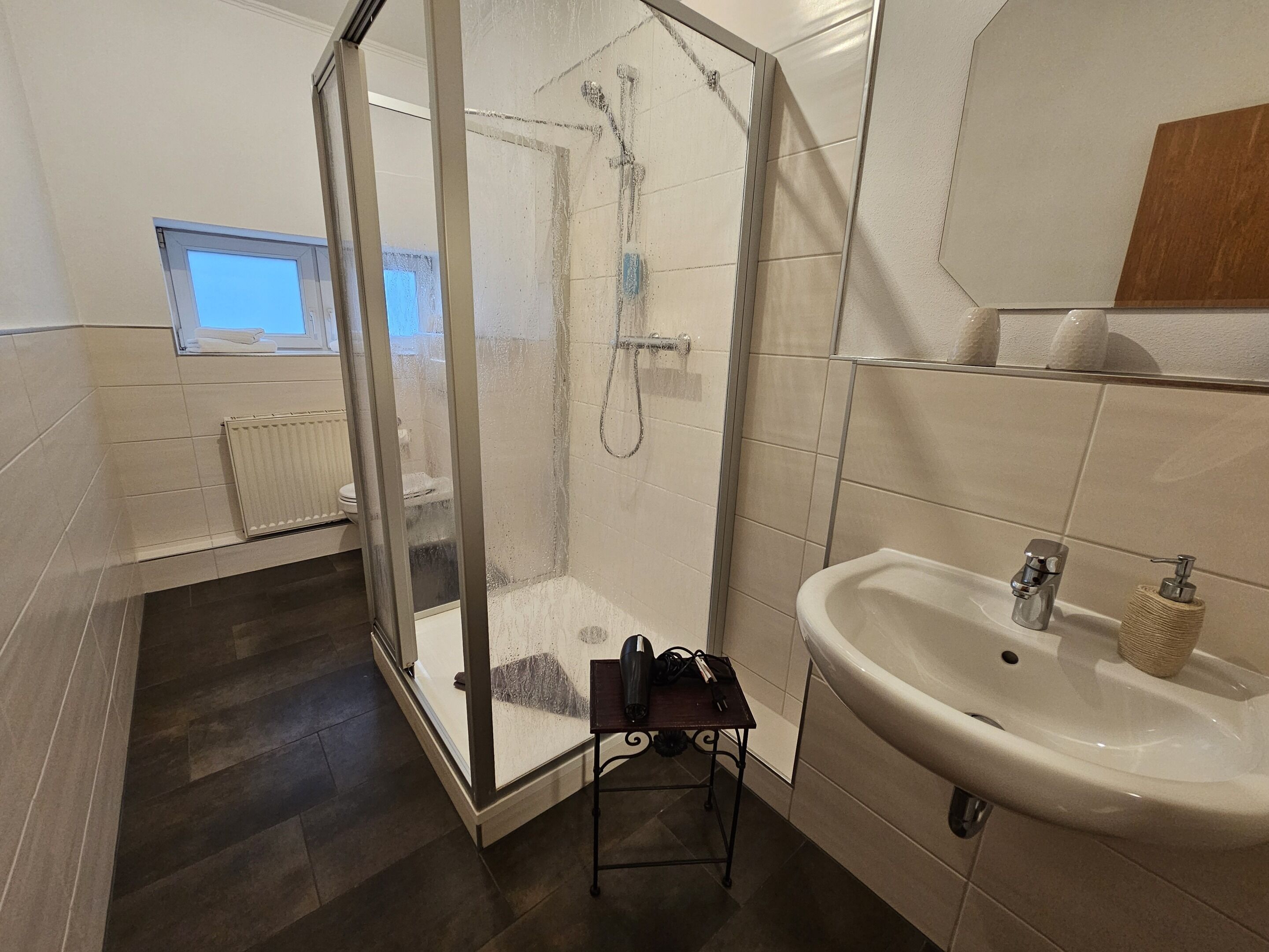 Un baño moderno con ducha envuelta en vidrio, un lavamanos blanco y una mesita con artículos de aseo.