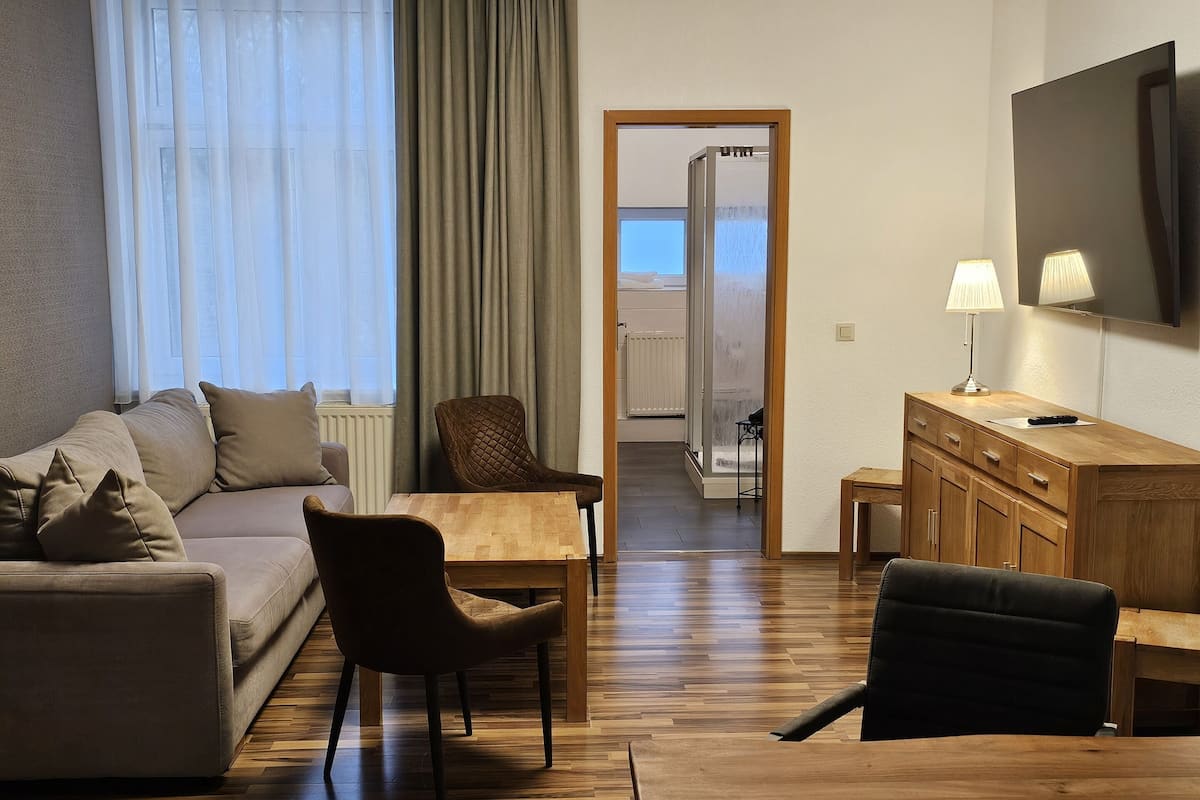 Ein modernes Wohnzimmer mit einer Couch, Sesseln, einem Holztisch, einem Fernseher und Blick durch eine Glastür.