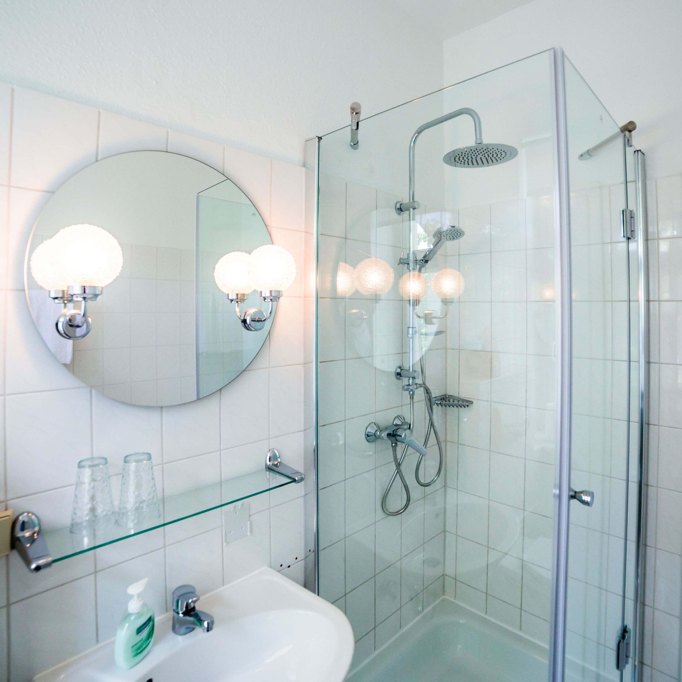 Un baño moderno con cabina de ducha de vidrio, un espejo redondo con apliques de pared y un estante de vidrio con artículos de aseo.