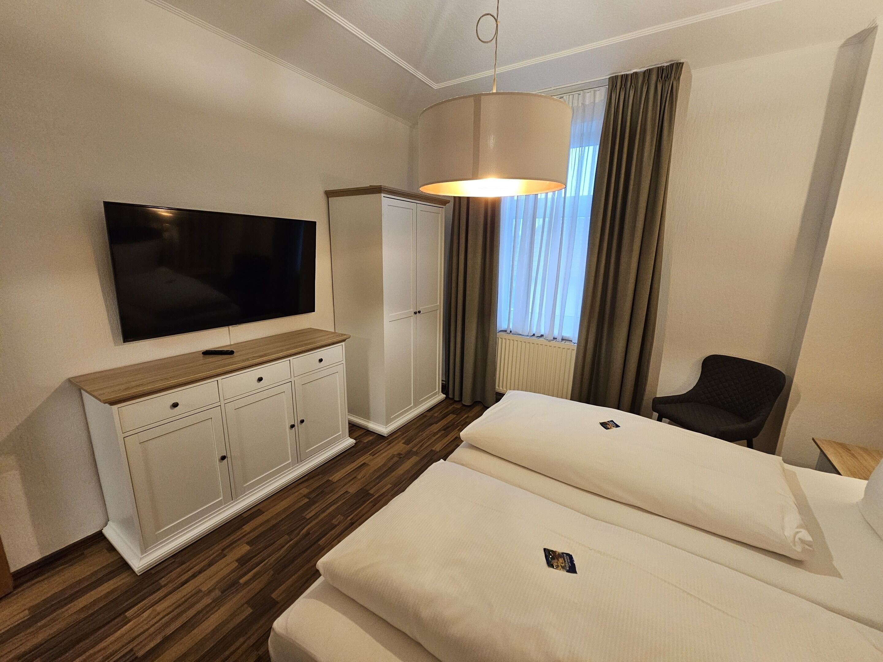 Habitación de hotel con televisión de pantalla plana, cómoda de madera, cama con dos almohadas, una silla y ventana con cortinas.