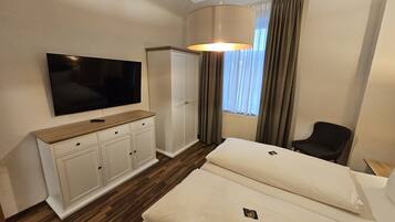 Habitación de hotel con televisión de pantalla plana, cómoda de madera, cama con dos almohadas, una silla y ventana con cortinas.