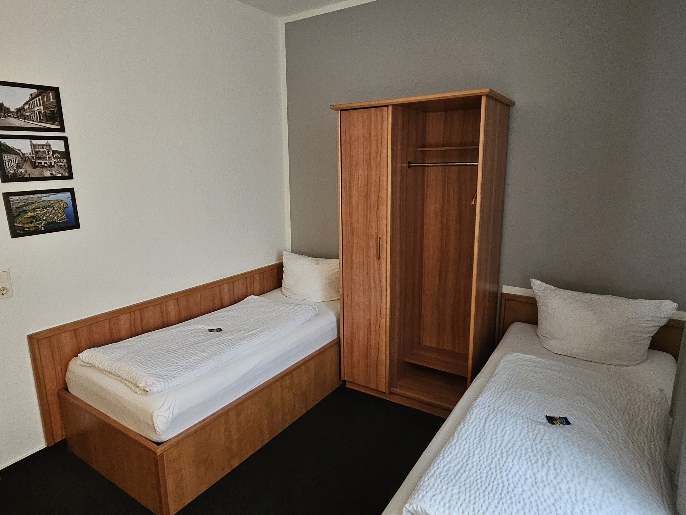 Una stanza modesta con due letti singoli, un armadio in legno e tre quadri appesi al muro.