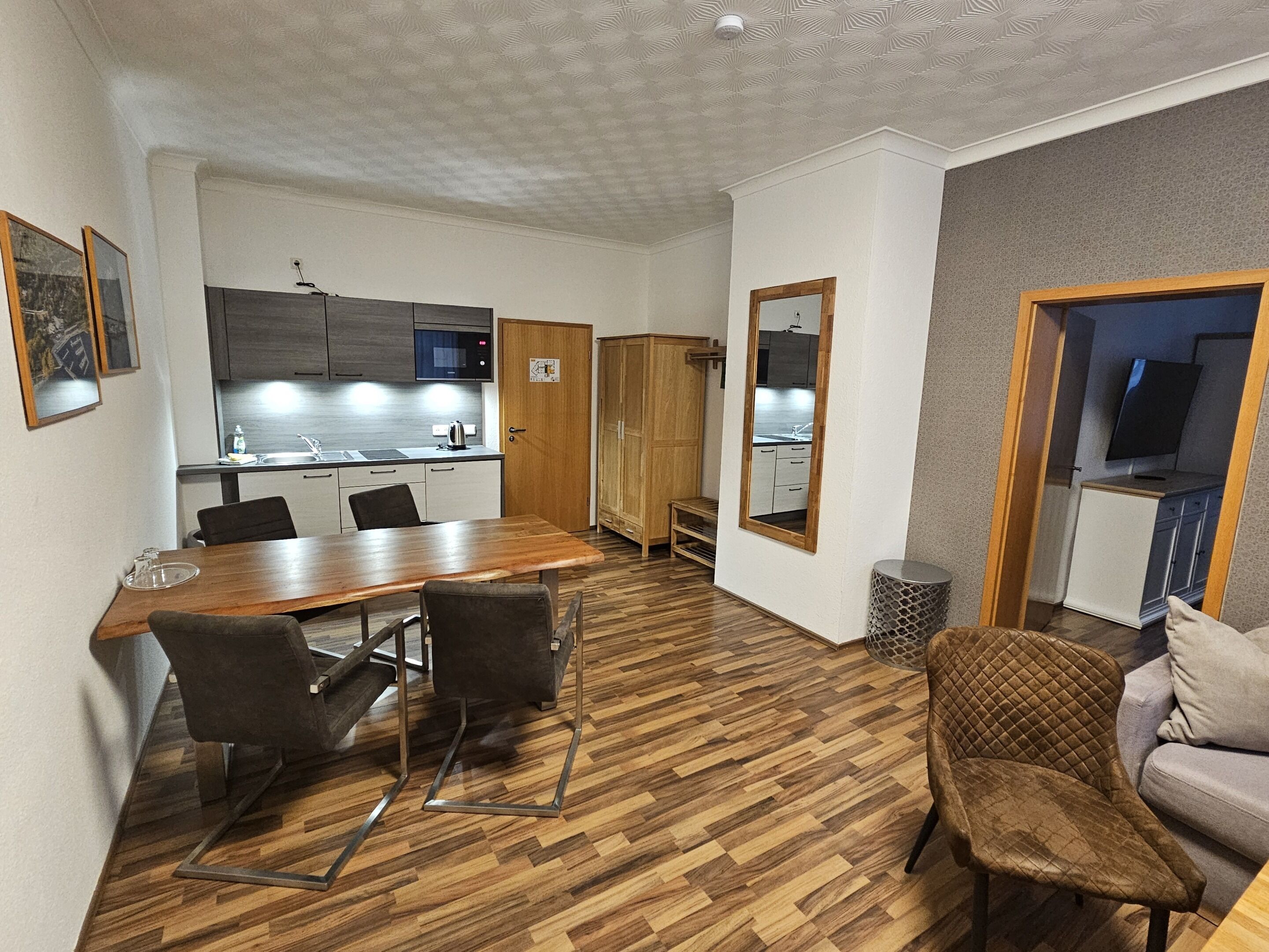 A modern, well-lit room with a wooden dining table, chairs, a sofa, and a TV.