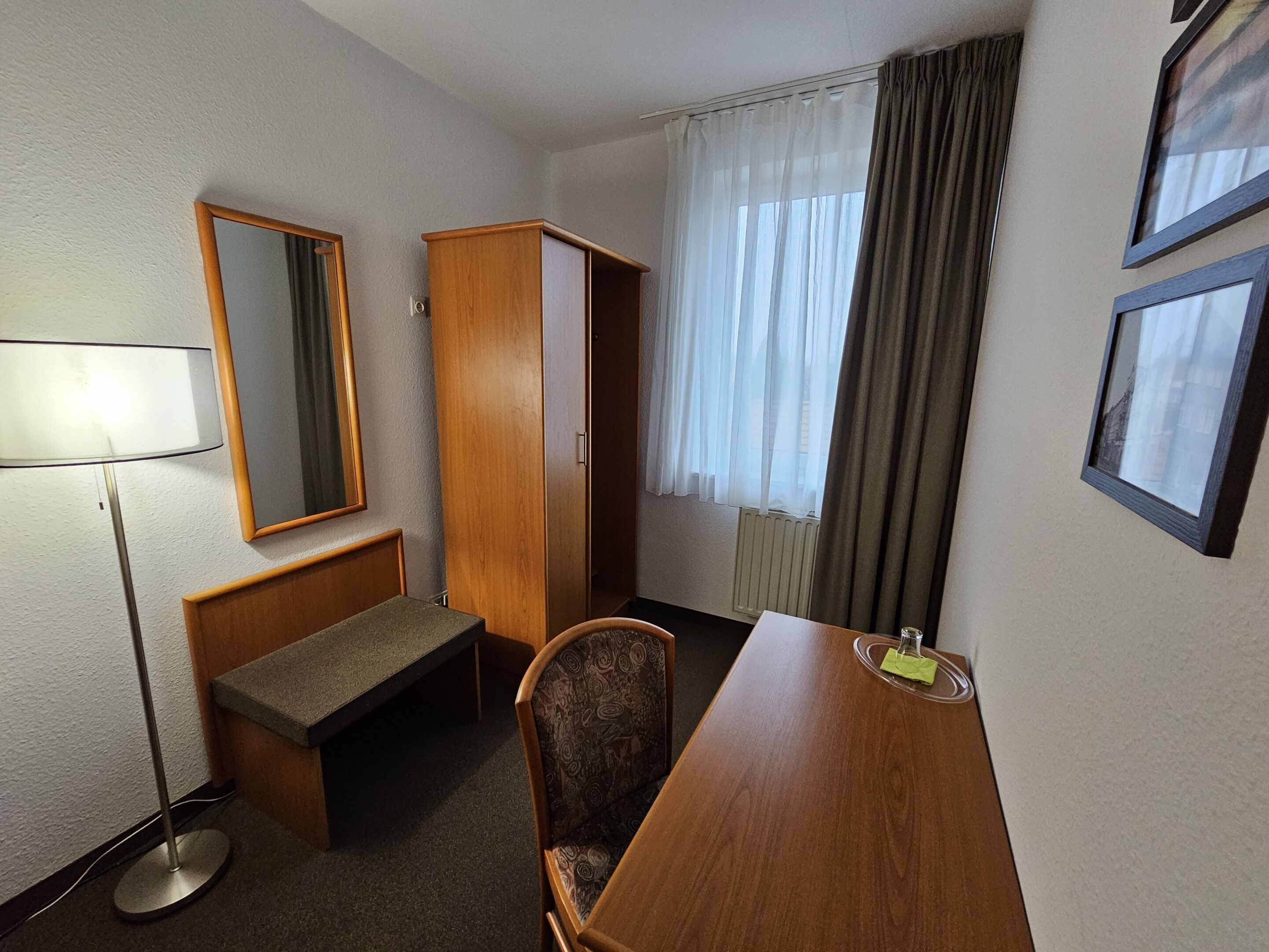 Hotellihuone, jossa on puinen työpöytä, tuoli, penkki, vaatekaappi, peili ja ikkuna, jossa on verhot.