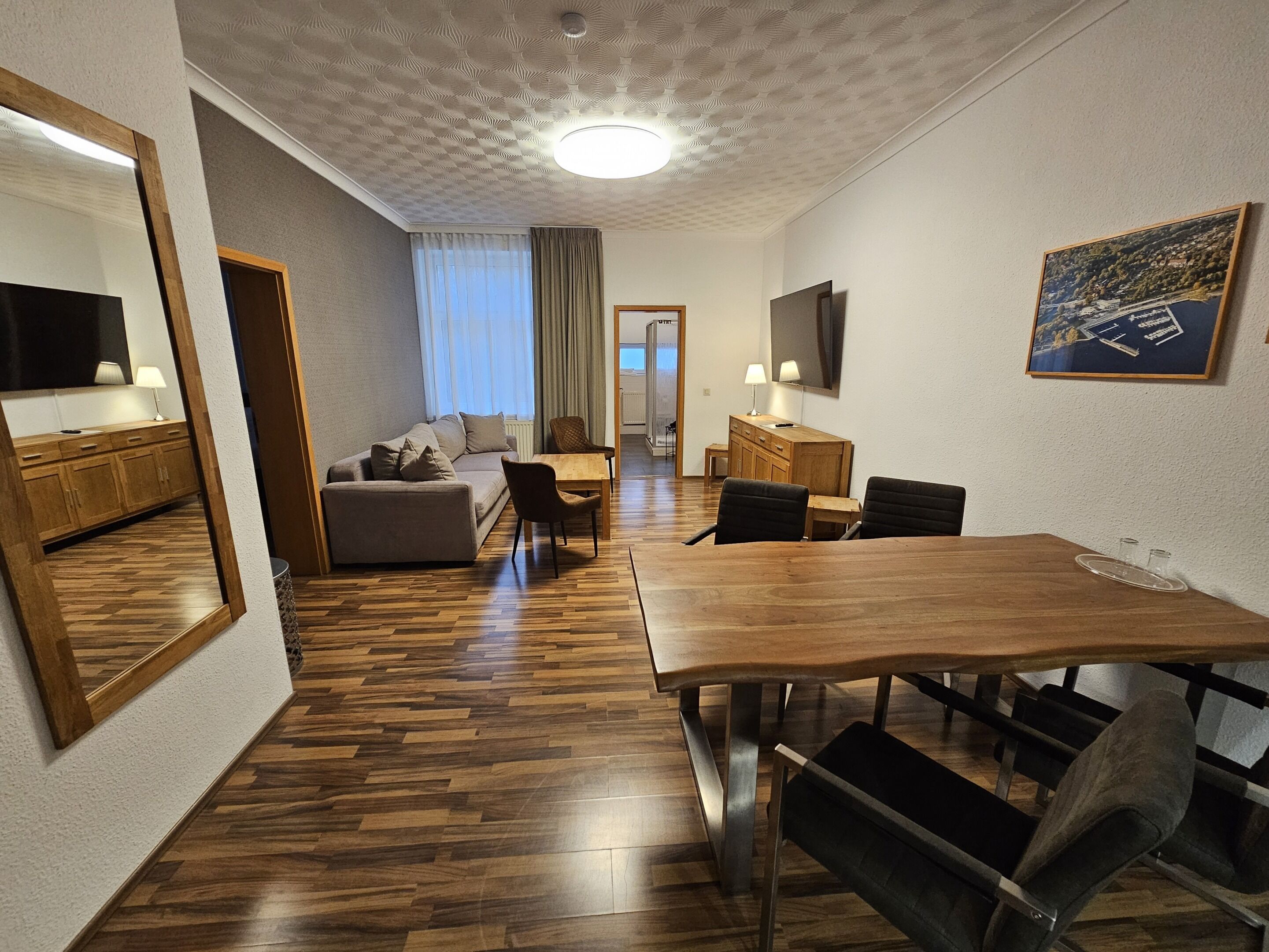 Habitación de hotel moderna con una mesa de comedor de madera, asientos cómodos, un sofá y un espejo grande.