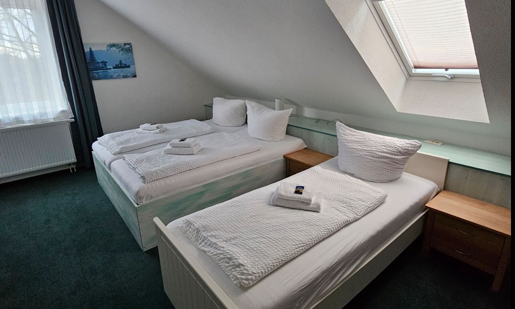 A twin room with two beds, each with white linens and towels, a small bedside table, a nightstand, and a window with blinds.