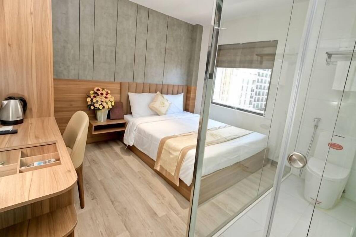 Une chambre d’hôtel comprenant un lit, un bureau, une chaise, une petite table, un vase avec des fleurs et un espace douche.
