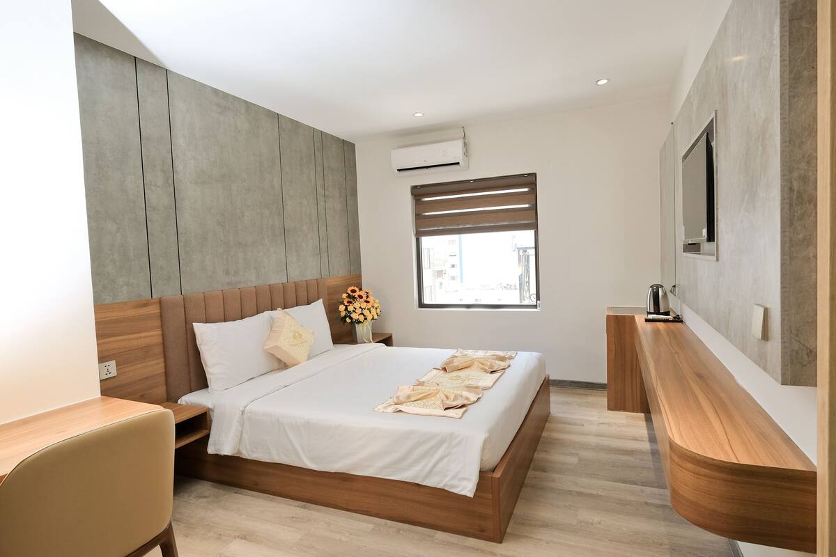 Une chambre moderne avec un lit en bois, une table de chevet, une commode, un téléviseur fixé au mur et une fenêtre avec des stores.