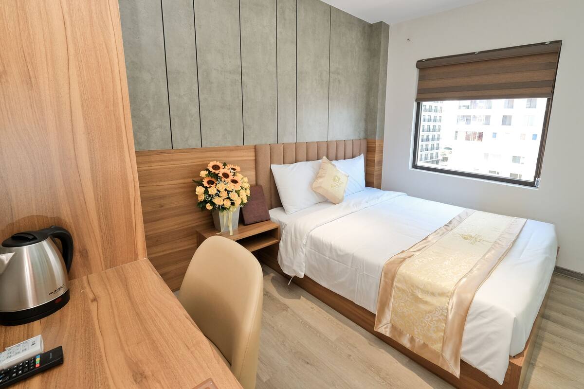 Une chambre d’hôtel avec un lit, un bureau en bois, une machine à café, une télécommande et une fenêtre donnant sur des immeubles.