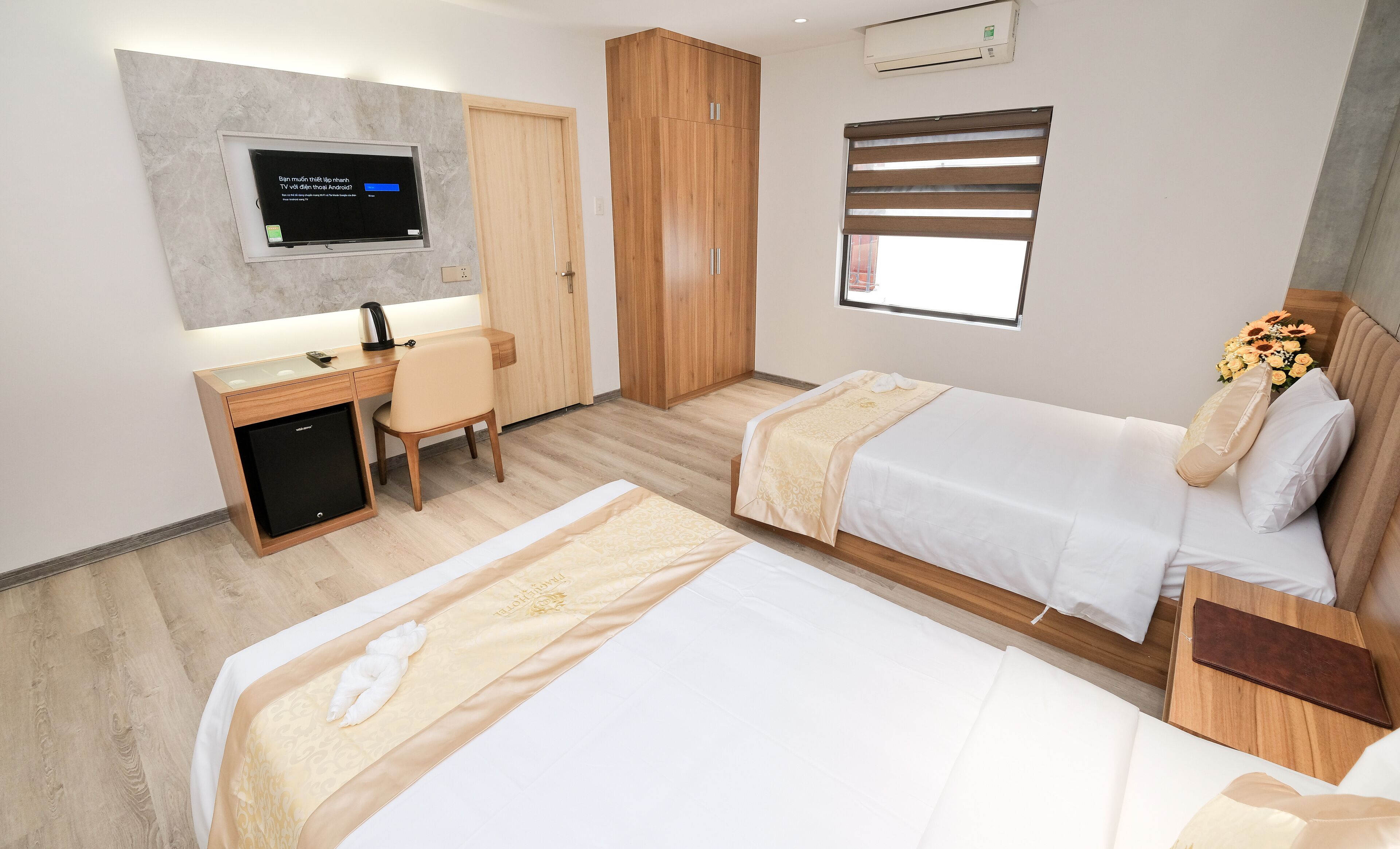 A hotel room with two beds, a desk, a chair, a TV mounted on the wall, and a window with blinds.
