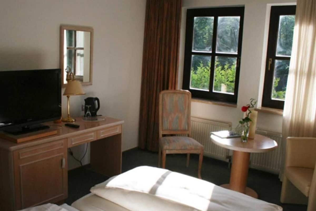 A hotel room with a bed, a desk with a TV, a chair, a small table with a vase, and a window with curtains.