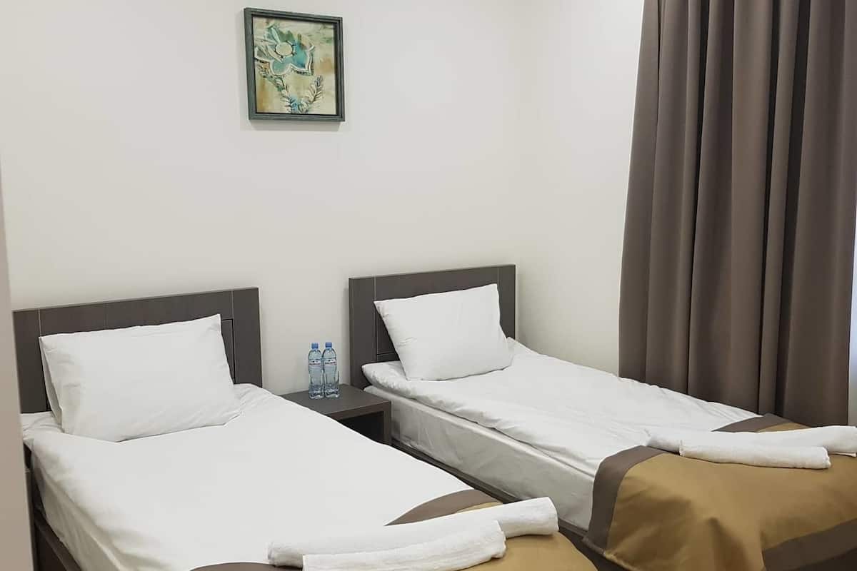 A hotel room with two single beds, a nightstand with water bottles, a framed picture on the wall, and a window with curtains.