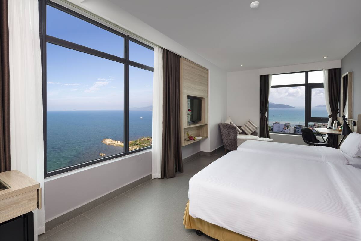 Une chambre d’hôtel dotée d’une grande fenêtre donnant sur la mer, d’un lit, d’un bureau et d’une chaise.