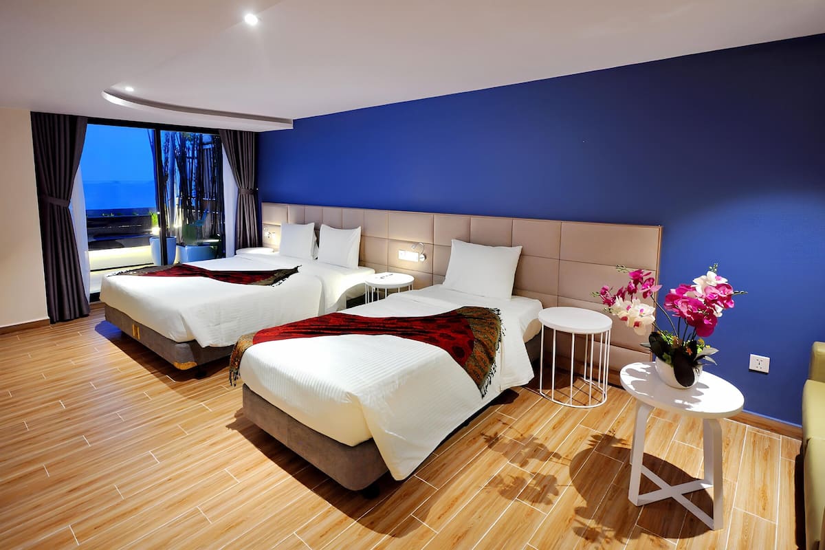 Une chambre d’hôtel moderne avec deux lits, une grande fenêtre avec des rideaux, une petite table ronde ornée d’un vase de fleurs et une vue sur l’extérieur.
